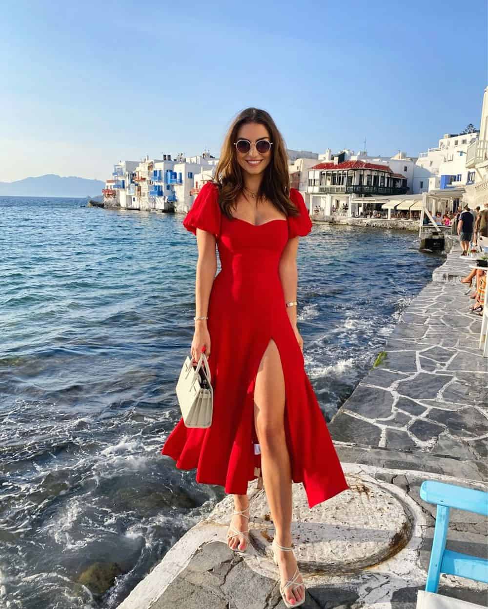image of a woman standing in front of the coast wearing a red midi dress and white heeled sandals 