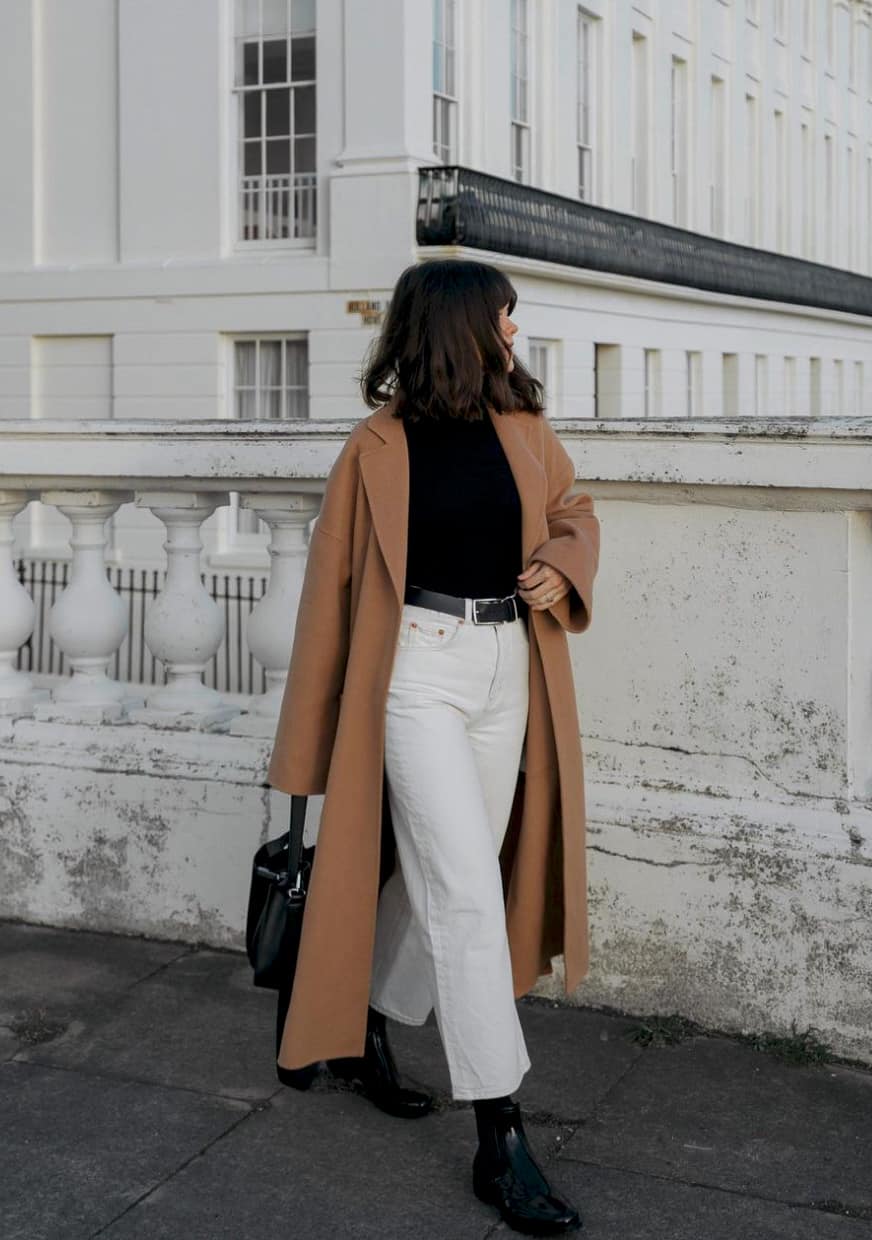 image of a woman walking down the sidewalk wearing a long wool camel coat, black turtleneck, white wide leg pants, and black boots