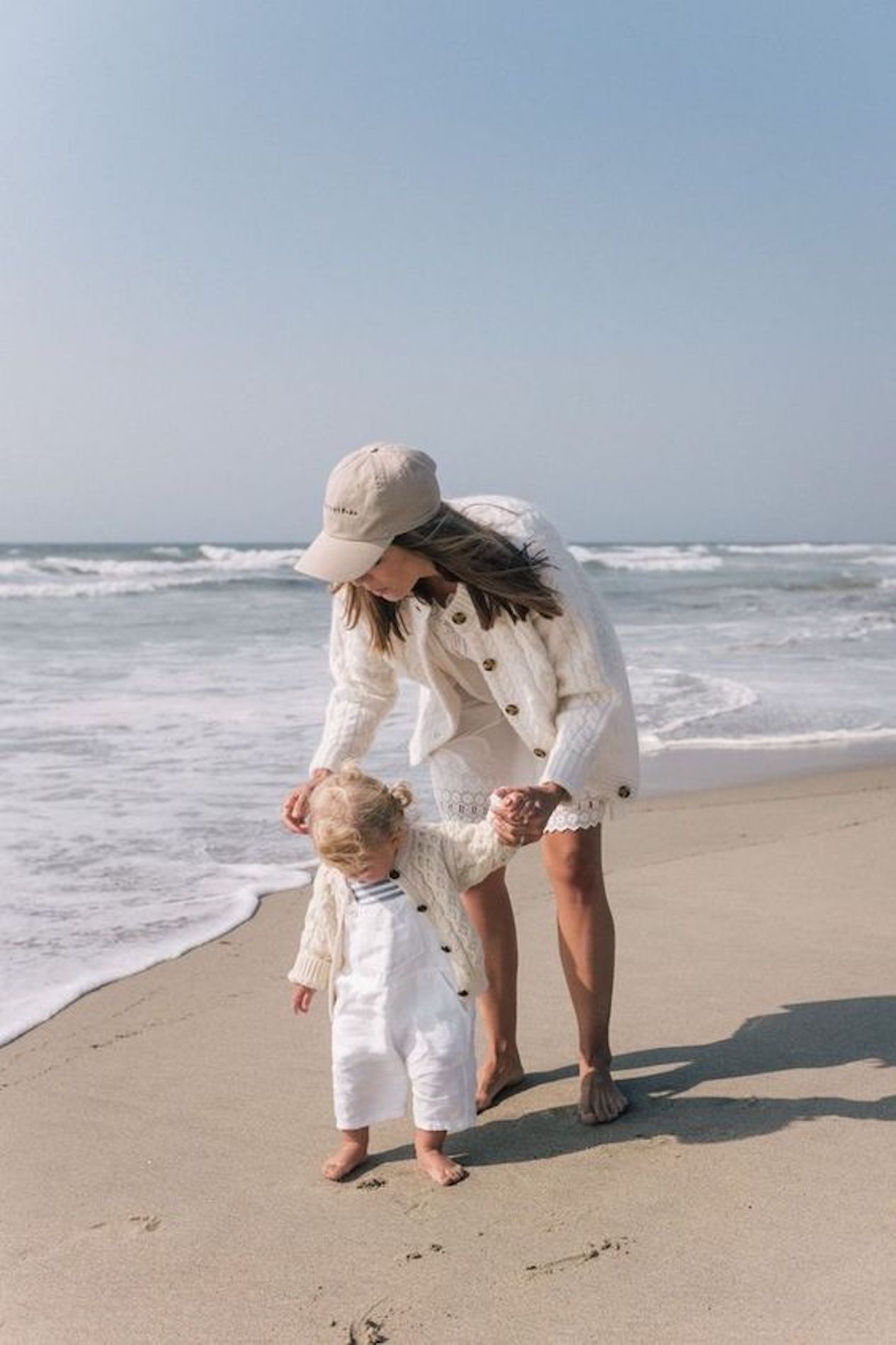 image of a mother and daughter at the beach with the ocean in the background wearing white clothing with ivory knit sweaters