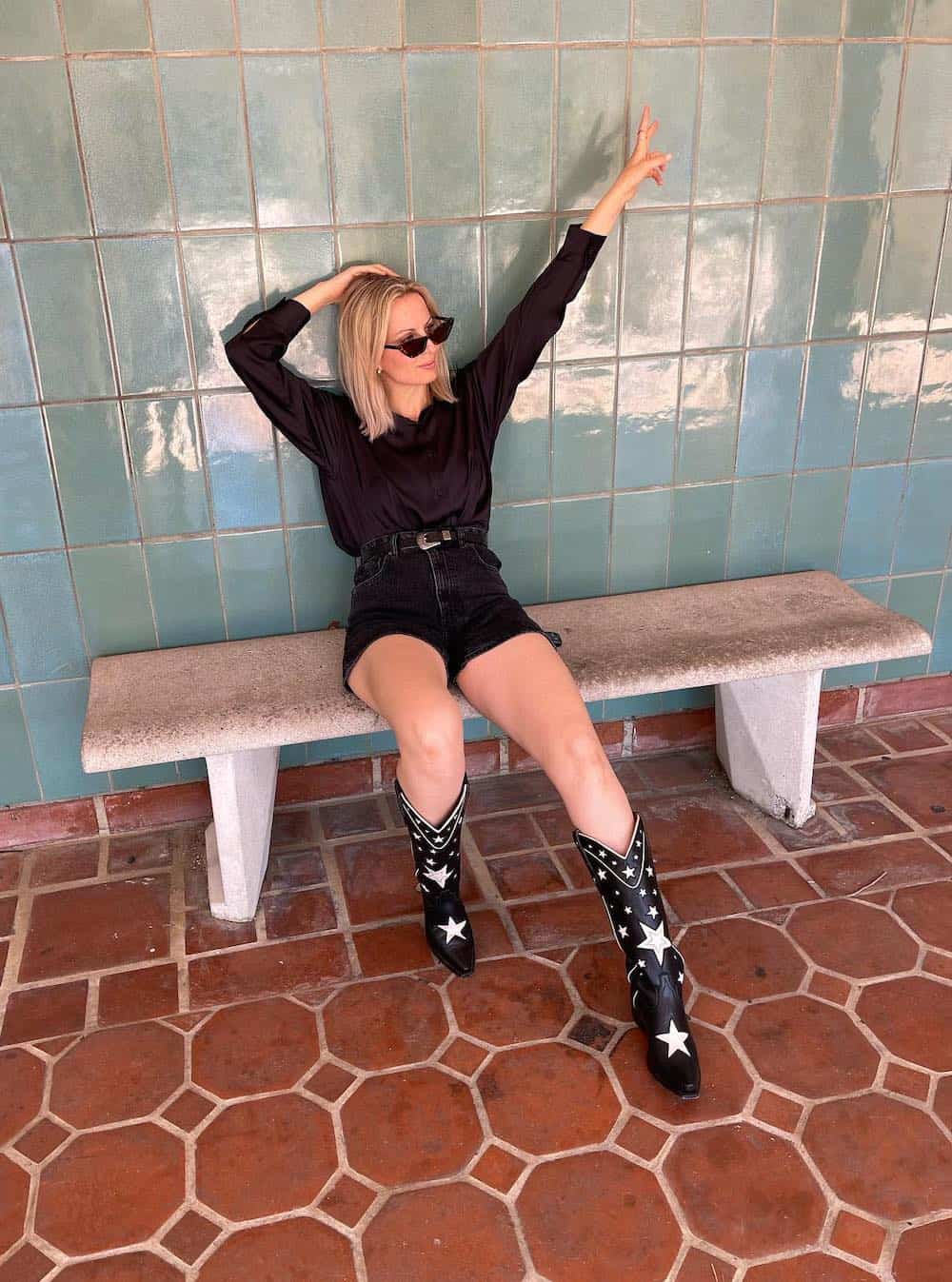 woman sitting on a bench wearing a black top, western belt, black denim shorts, and black and white cowboy boots