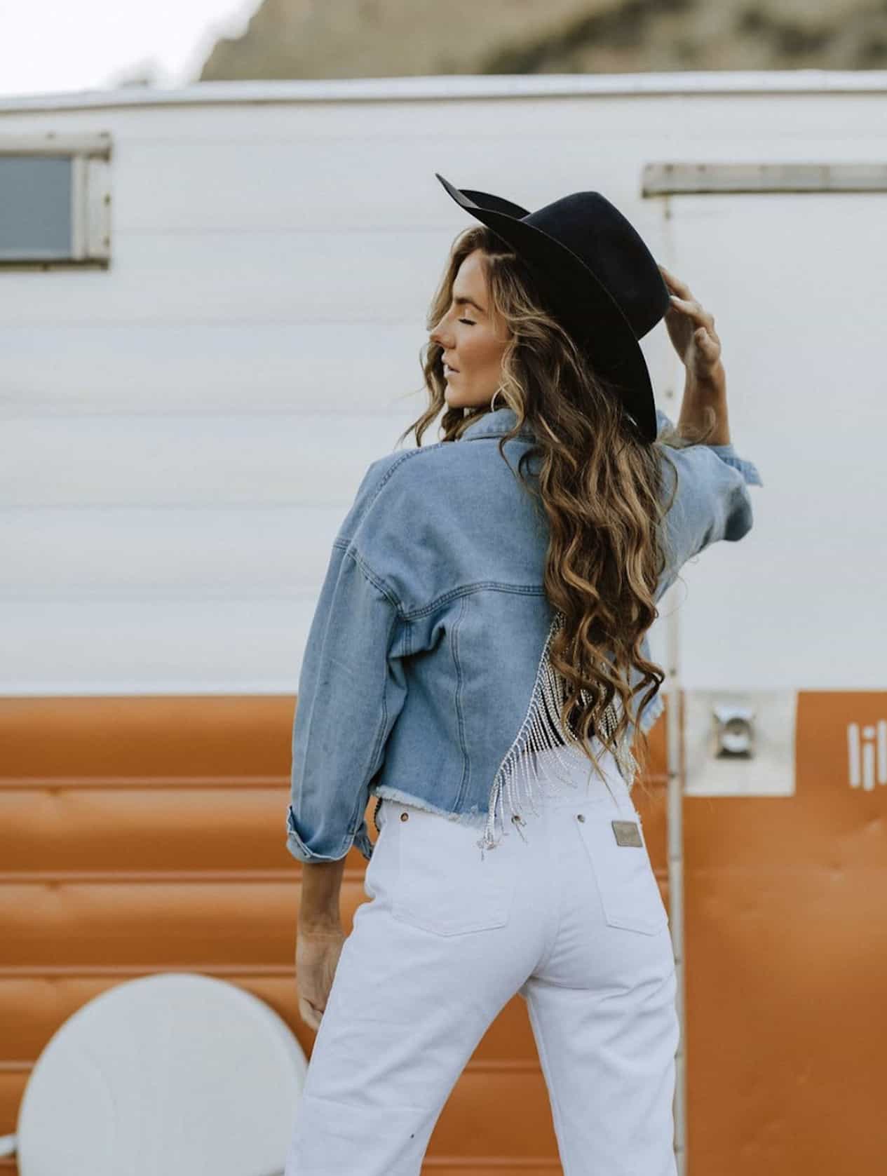 the backside of a woman standing in front of a trailer wearing white wrangler jeans, a denim jacket, and a black cowboy hat