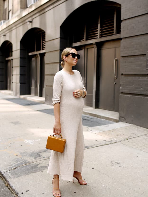 image of a pregnant woman standing on a sidewalk wearing a long cream colored sweater dress and dark sunglasses
