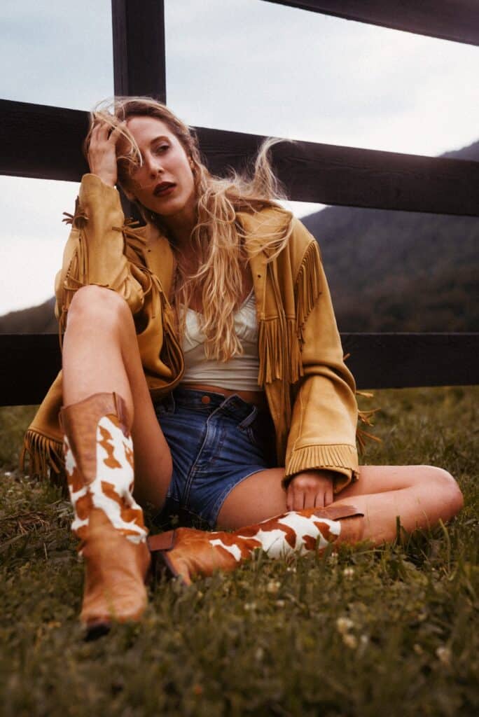 a woman sitting on the grass in front of a fence wearing a brown leather fringe jacket and cowhide western boots