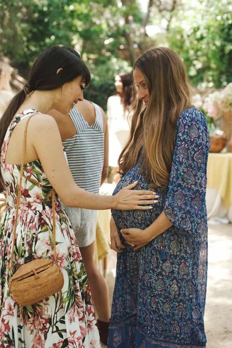 image of two woman at a garden party wearing dresses, one pregnant, and the other reaching out to touch the pregnant woman's belly