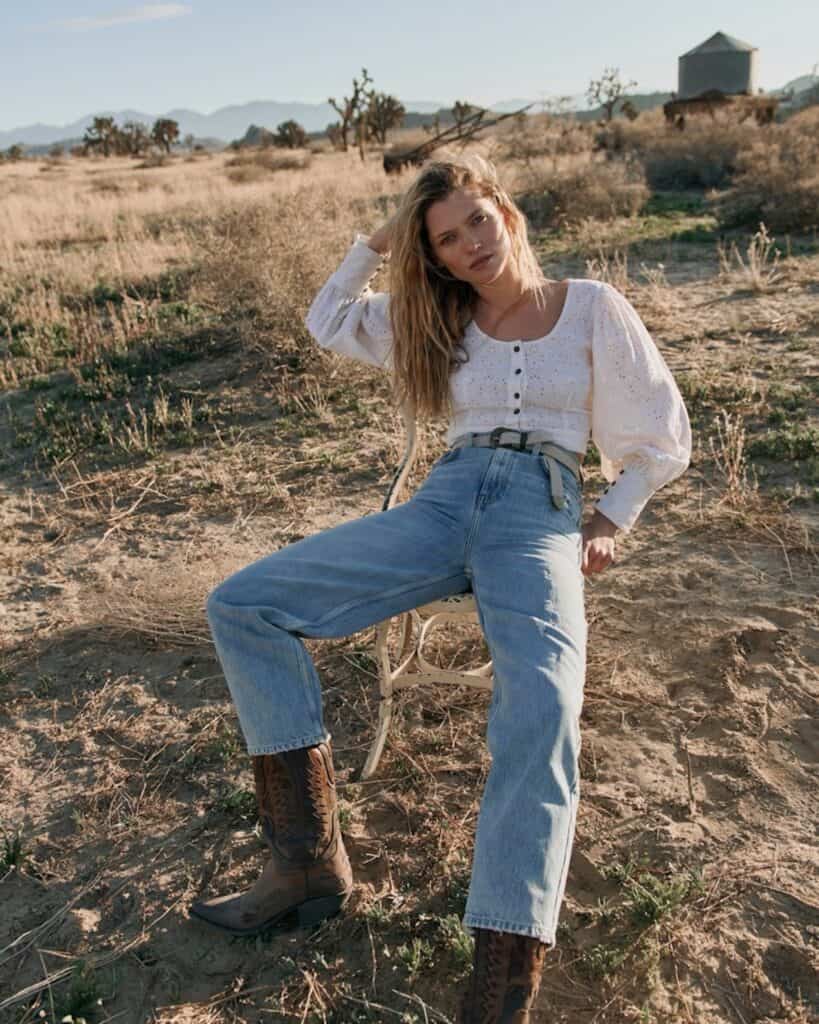 an image of a woman sitting in a chair in a field wearing a white blouse, jeans, and cowboy boots