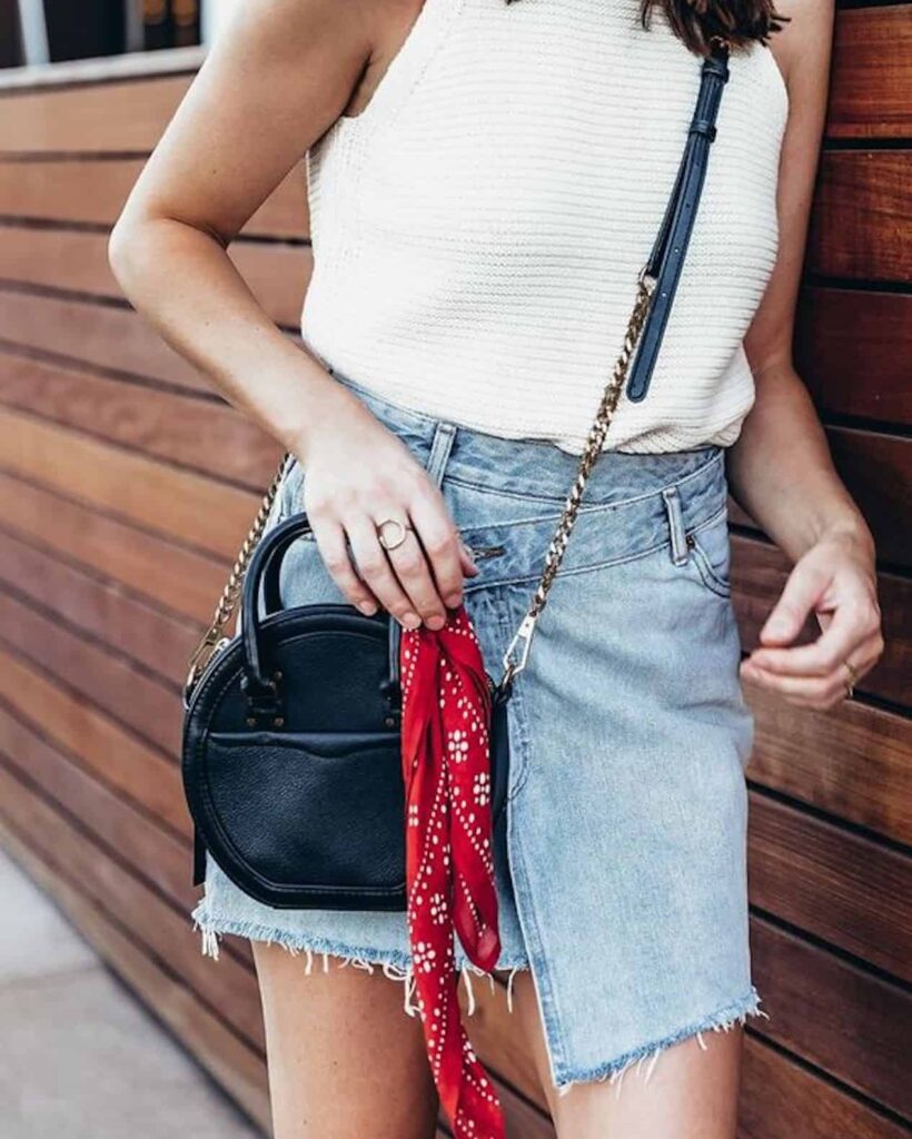 an image of a woman standing against a brick wall wearing a denim skirt, white top, and a black purse with a red bandana tied around it