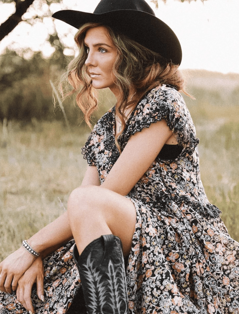 an image of a beautiful woman sitting in a field wearing a black cowboy hat, floral dress, and black cowboy boots
