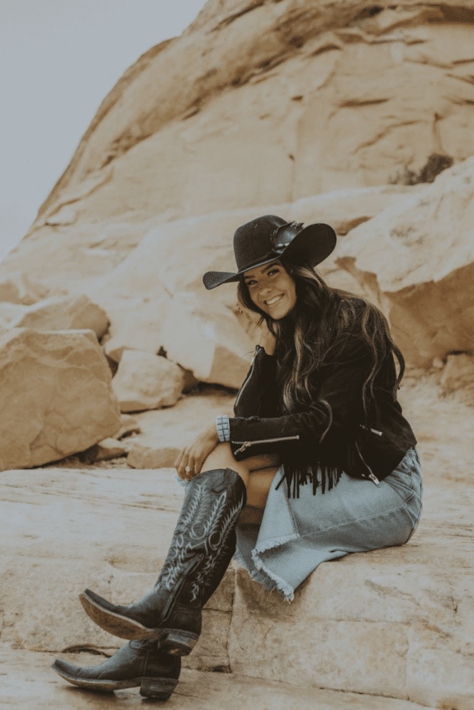a woman sitting in a rocky terrain wearing a black leather fringe jacket, a jean skirt, and black cowboy boots