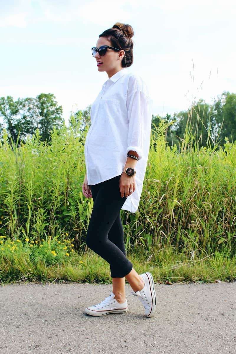 image of a pregnant woman standing in front of a grassy field wearing black leggings, sneakers, and an oversized white button up shirt