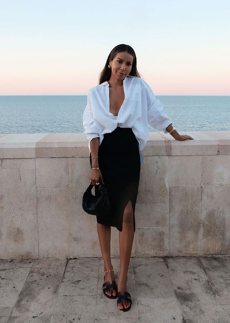 image of a woman standing against a ledge with the ocean in the background wearing a white button up shirt and a black skirt with black sandals