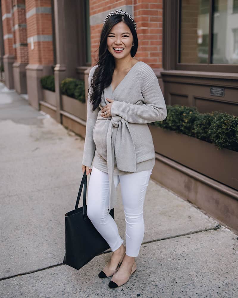 image of a beautiful pregnant woman standing on a sidewalk wearing a beige wrap sweater, white jeans, and a black purse