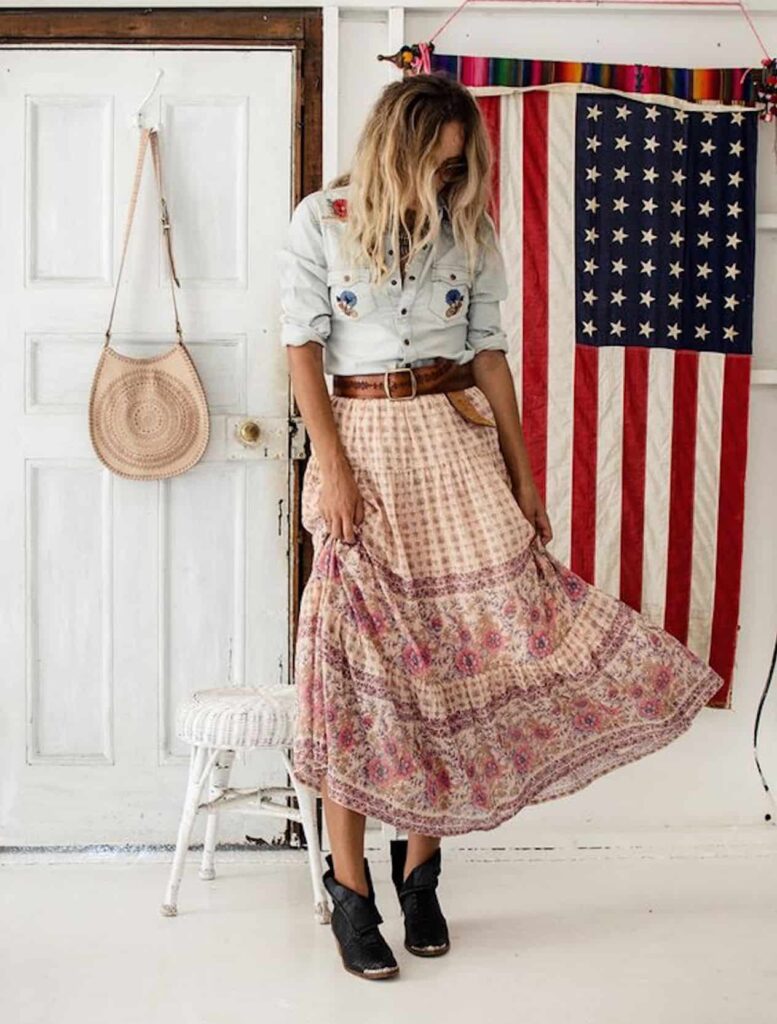 an image of a woman standing in a white room wearing a floral skirt and a denim button up shirt with an American flag in the background