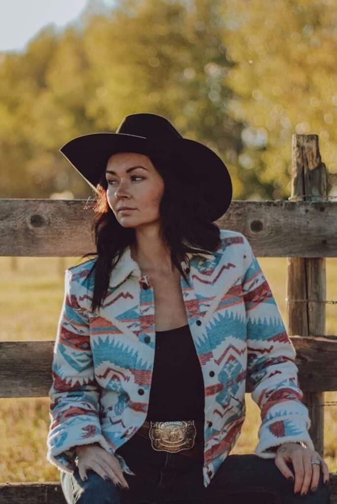 a woman sitting in front of a wood fence wearing western attire and an aztec print sherpa jacket