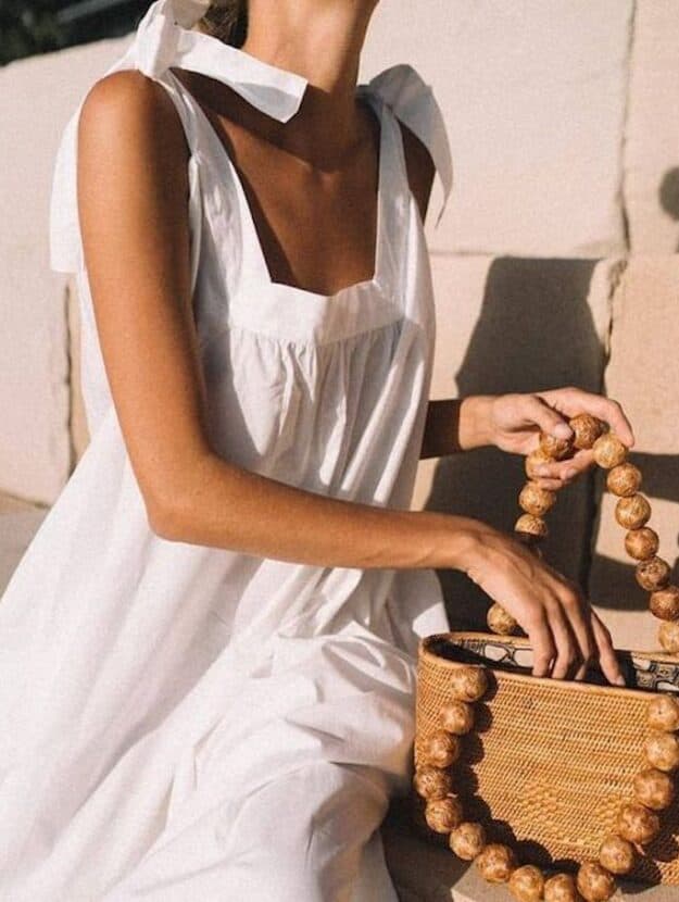 image of a woman reaching into a brown woven tote bag wearing a flowy white sun dress with tie shoulders