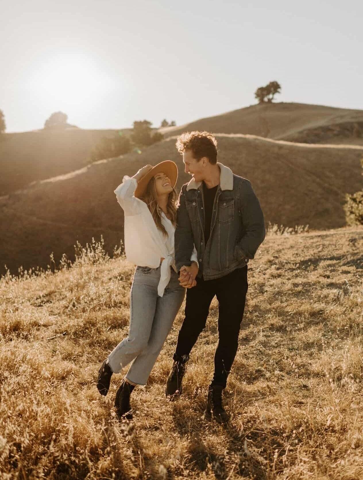 Young man and young woman hold hands and look at each other lovingly outside on a hill.