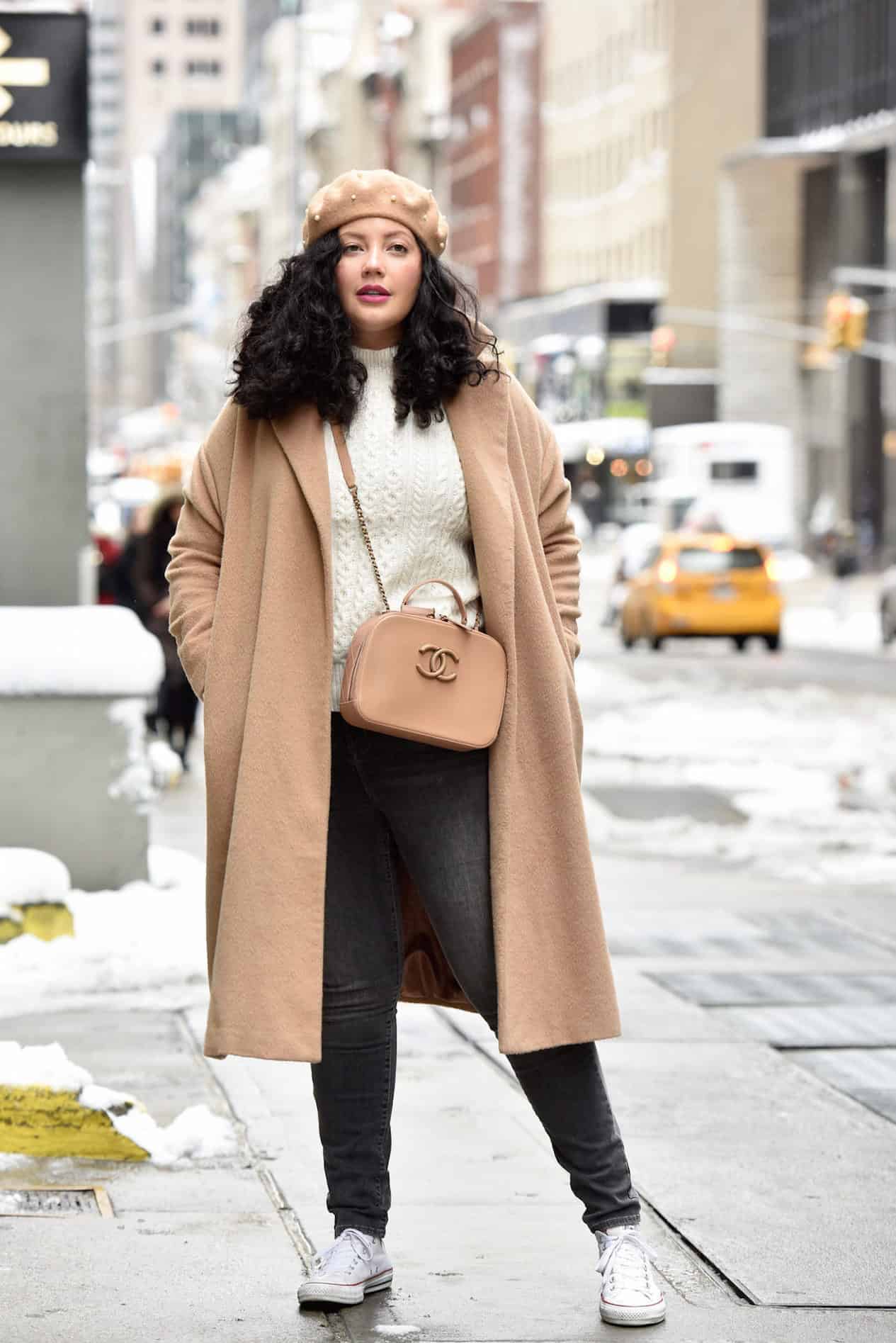 Young woman in black skinny jeans and camel colored coat poses outside in the city.