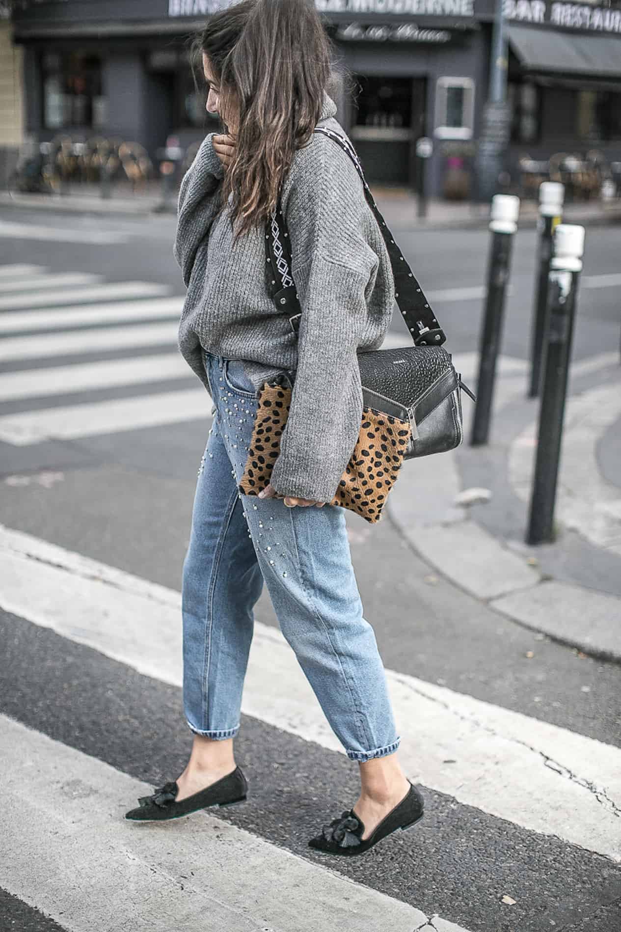 Young woman in grey sweater and embellished boyfriend jeans walks in the street.