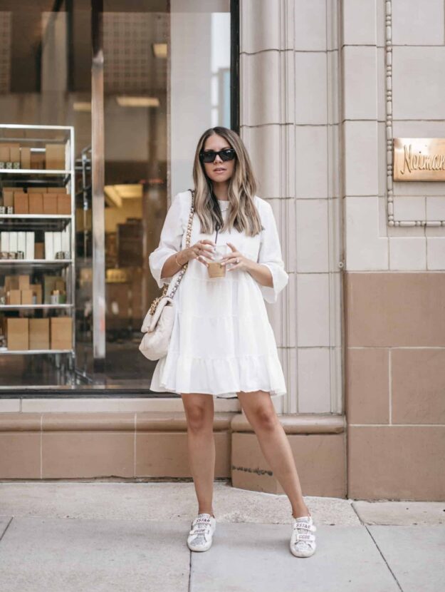 Young women in short white dress and sneakers poses in front of a Neiman Marcus.