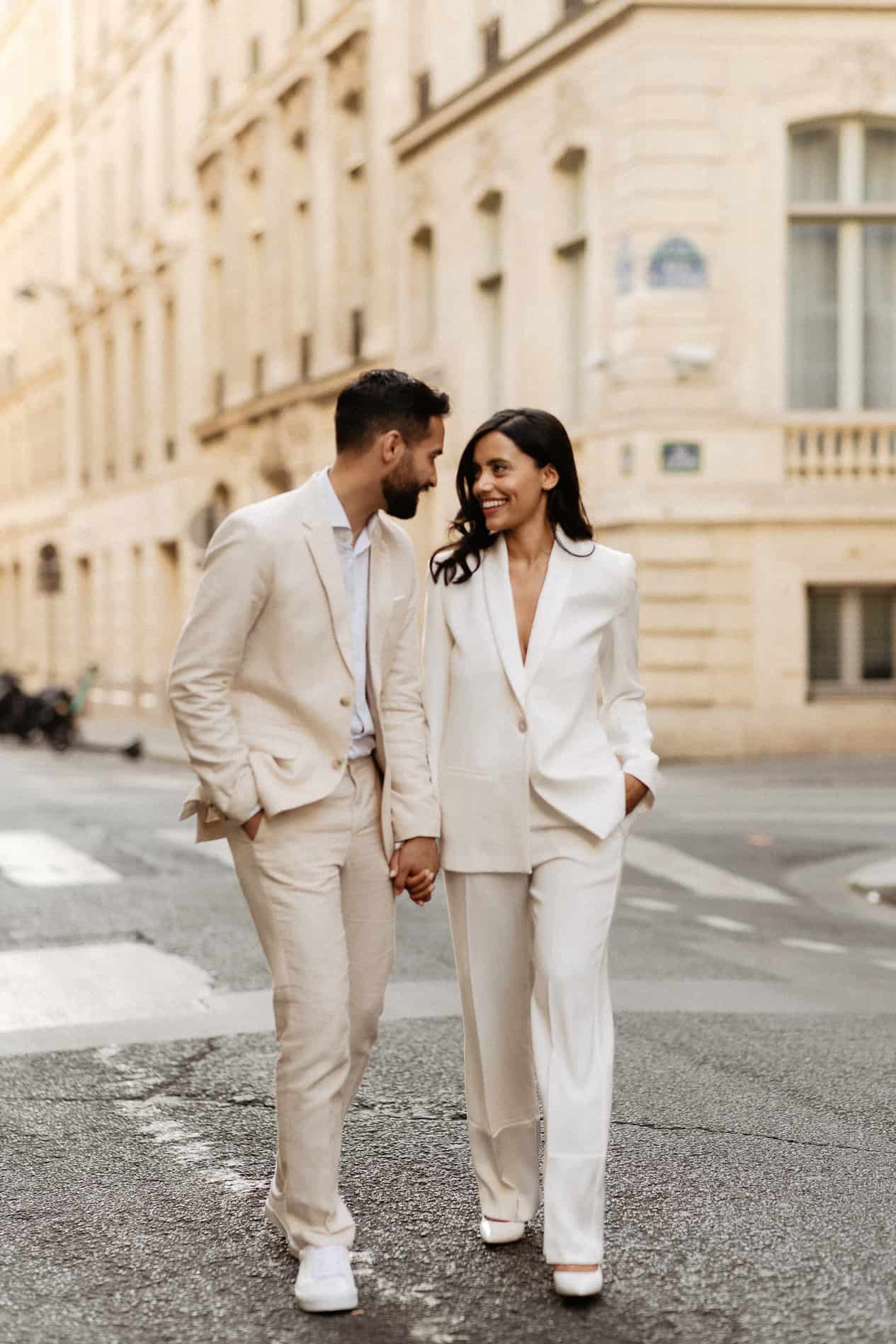 Young man and young woman walk in the street holding hands wearing neutral-colored suits.