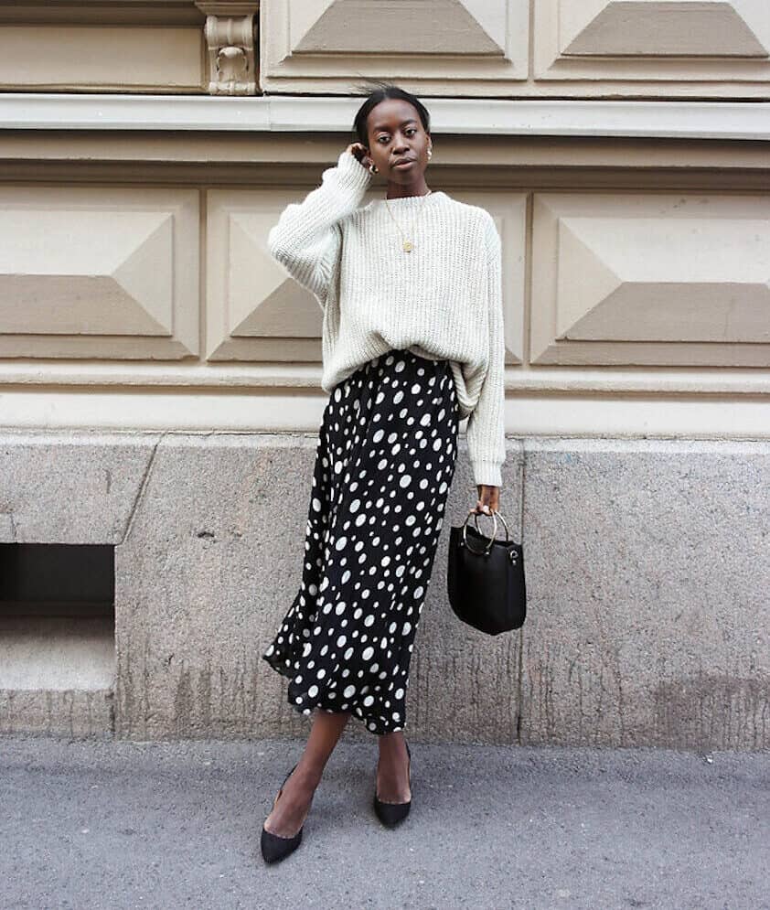 Woman wearing a black and white slip skirt and a white sweater poses in front of aa city building