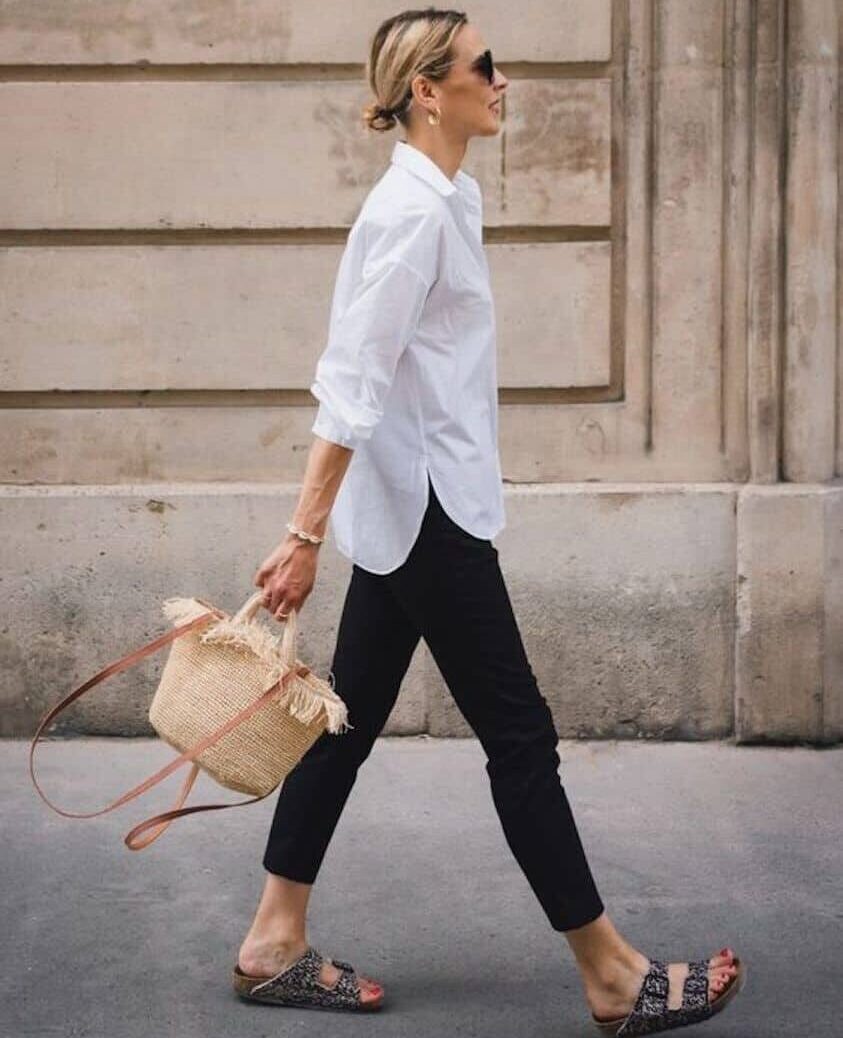 Woman wearing black skinny jeans, a white button down and Birkenstock sandals walks through the city.