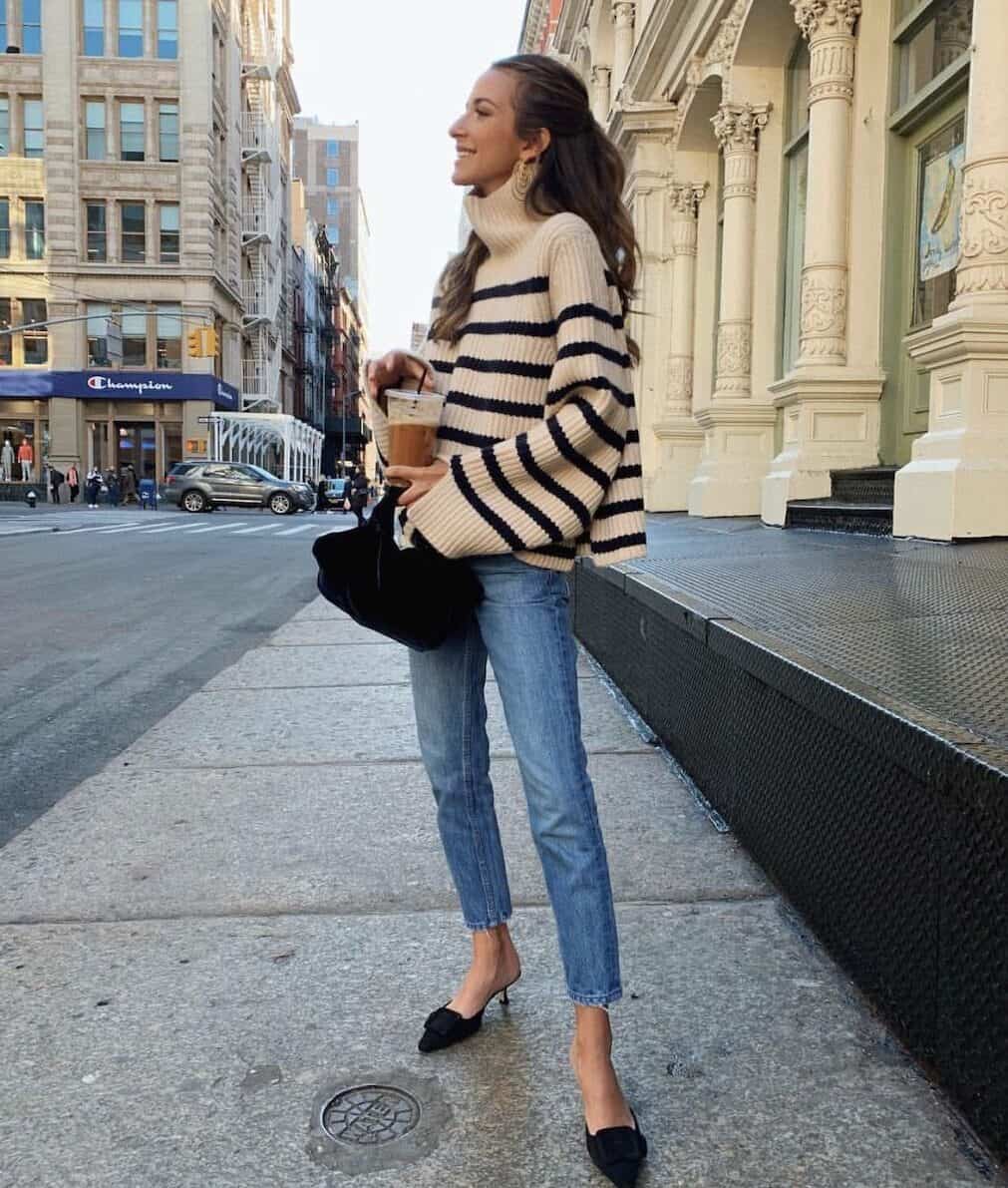 Woman wearing a black and cream stripped sweater, jeans, and black kitten heels drinks an iced coffee.