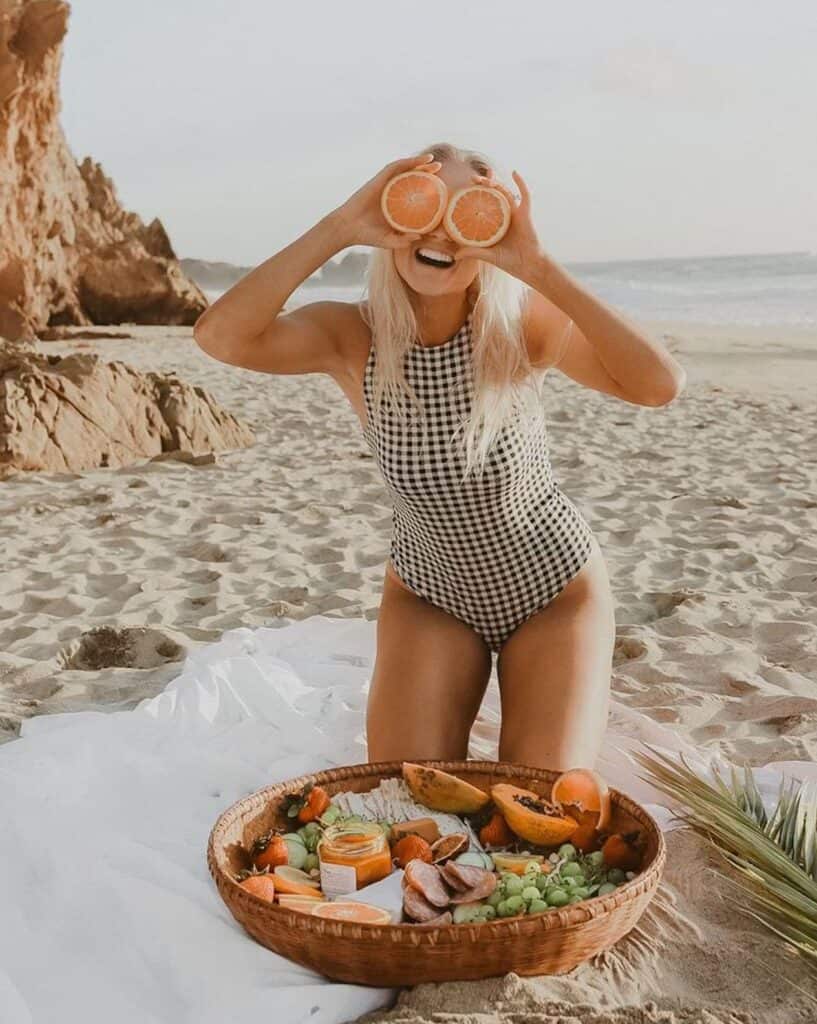 a woman in a swimsuit on the beach having a picnic holding an orange over her eyes