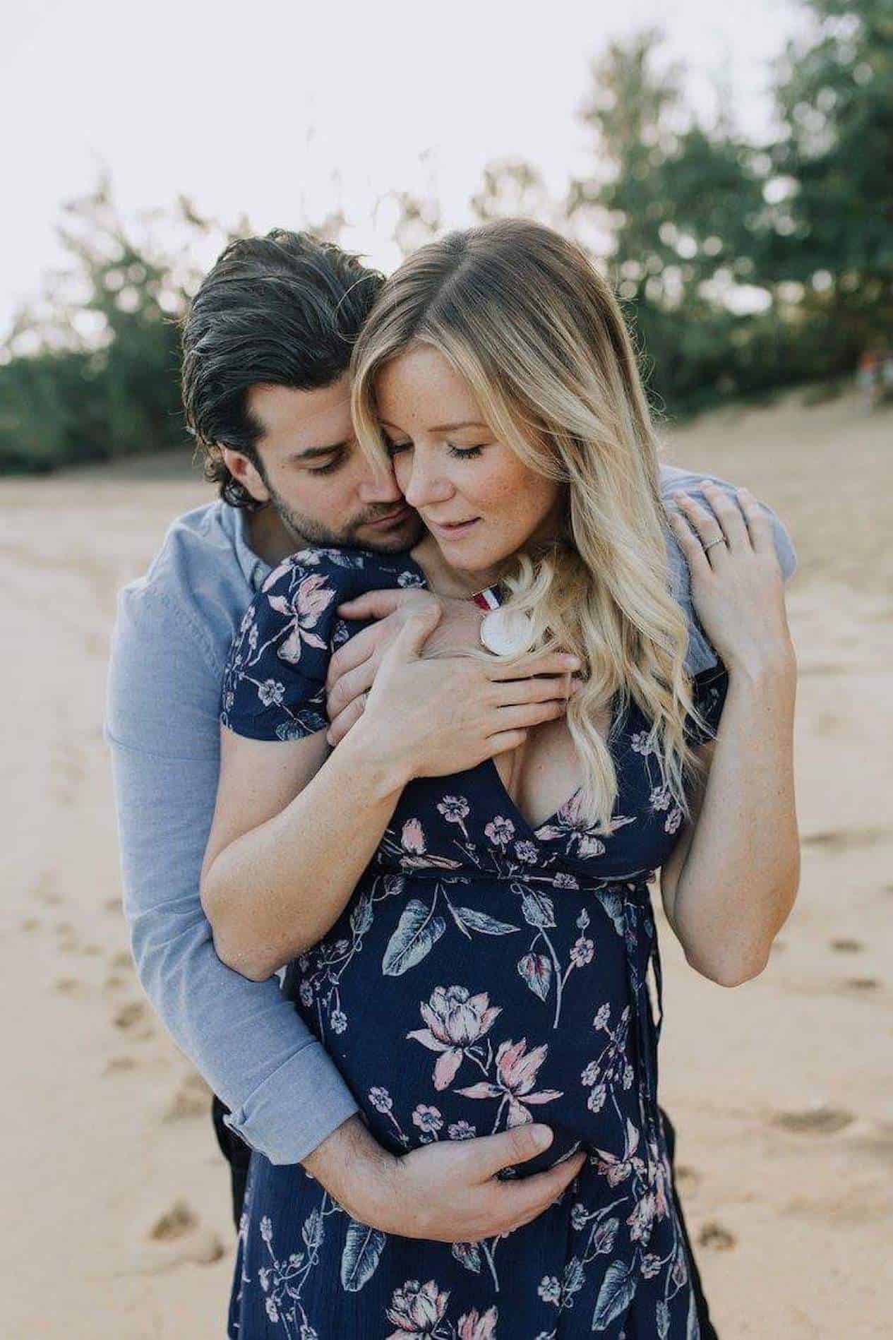 image of a man standing behind a pregnant woman on the beach giving her a hug, she is pregnant and wearing a blue floral dress