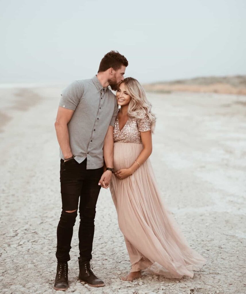 image of a man and woman standing on a wide beach, she is wearing a pink gown and is pregnant, and he is in black jeans and a grey shirt