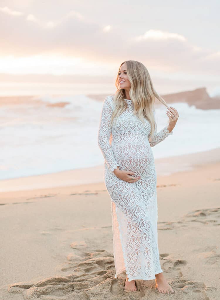 pretty pregnant woman standing on a beach at sunrise in a long white lace grown