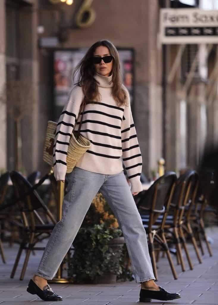 A woman wearing jeans, black loafers, a white and black striped turtle neck and a straw handbag walks near a cafe.