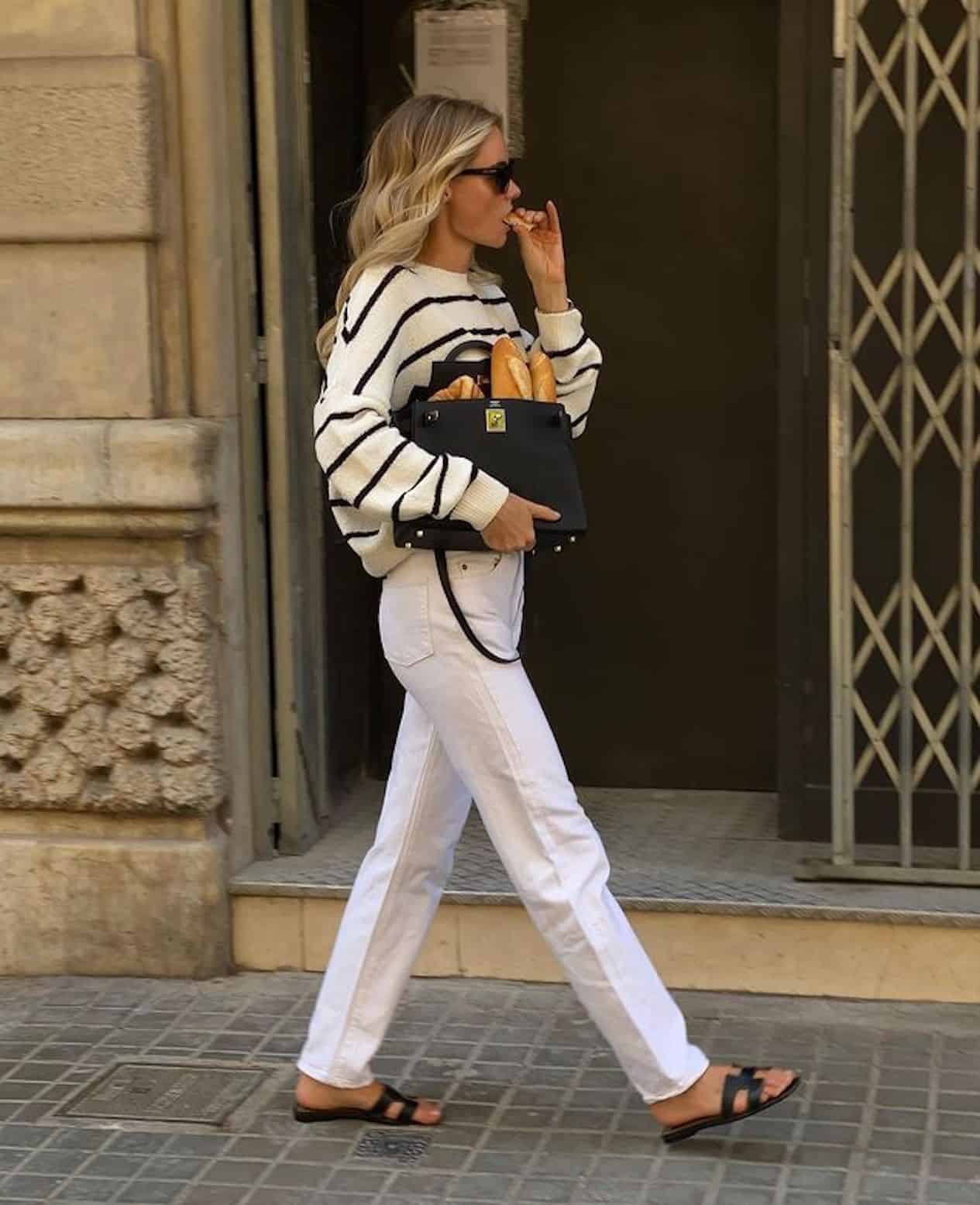 Woman wearing white jeans, black sandals and a black and white striped sweater walks through the city eating bread.
