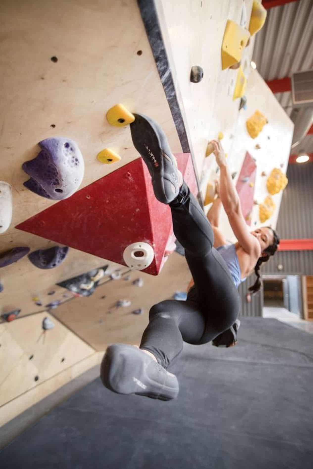 Woman indoor rock climbing in black leggings and a grey tank top.