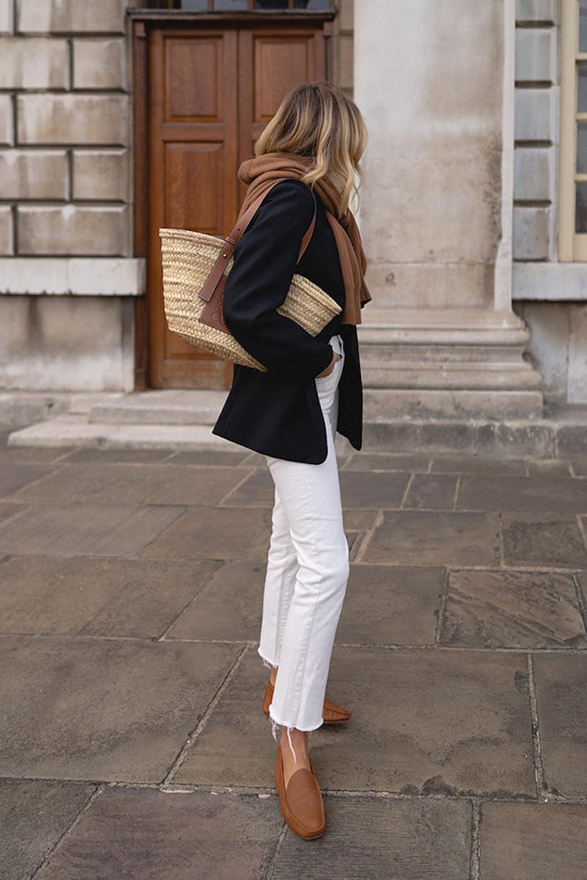 Woman wearing white jeans, tan loafers, a black blazer and a brown scarf walks through the city.