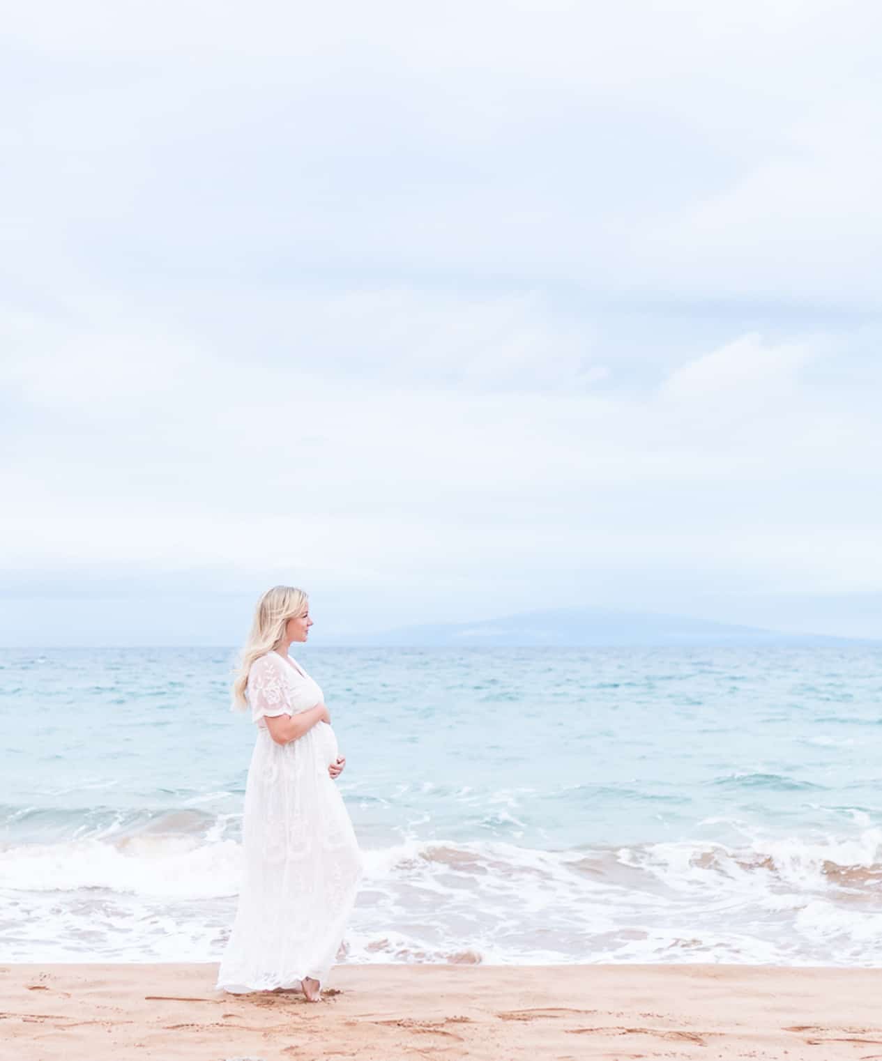 image of a woman from a distance standing on a beach in front of the ocean wearing a white flowy dress embracing her pregnant belly