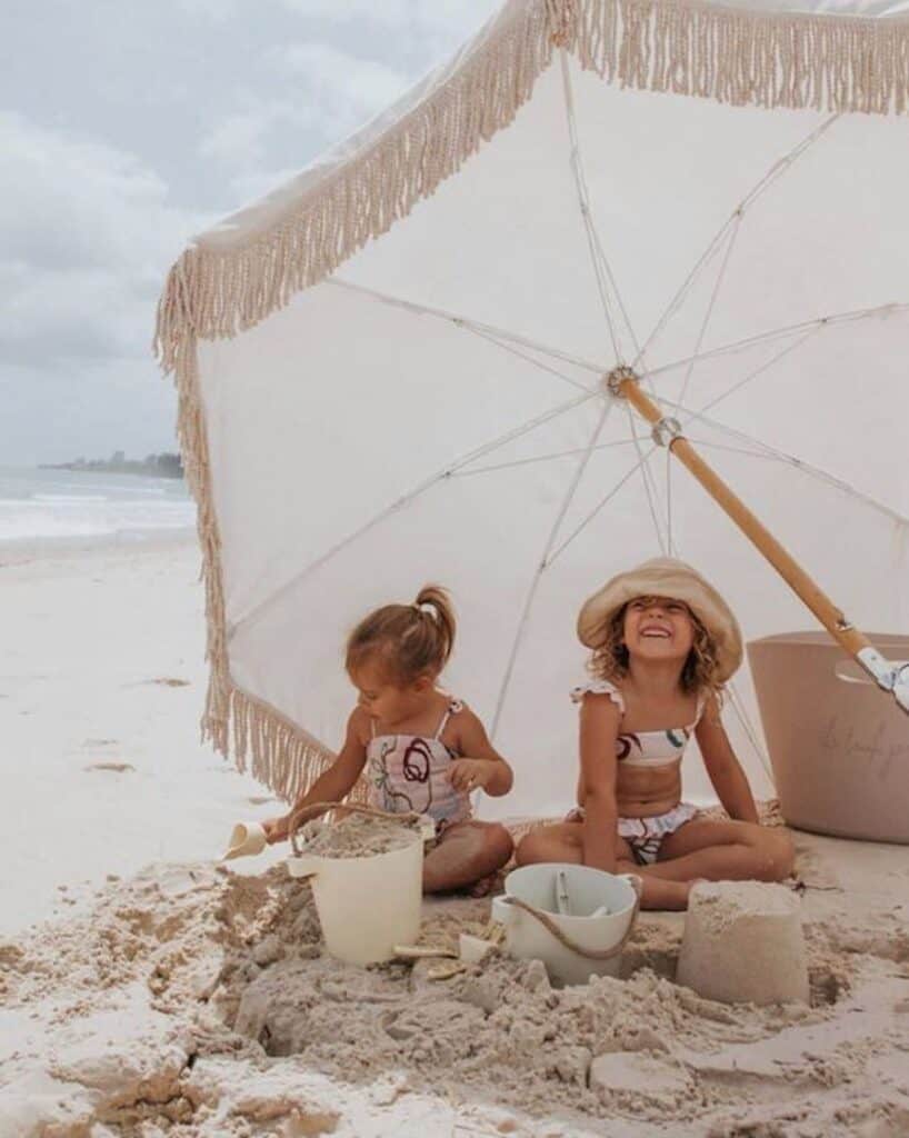 an image of two young girls sitting on the beach underneath and umbrella playing in the sand