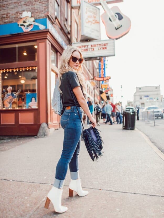 Woman walking down Broadway street in Nashville wearing jeans, white booties, a graphic tee, a fringe purse and sunglasses.