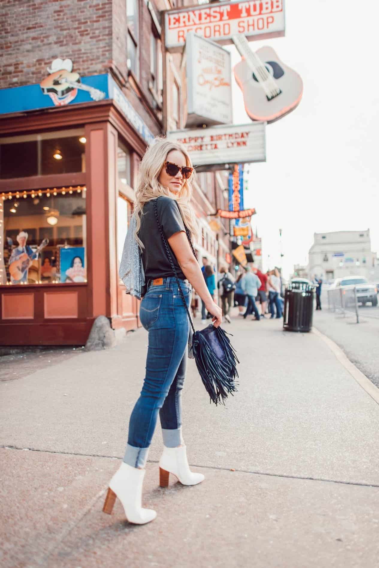 Phụ nữ đi bộ trên đường phố Broadway ở Nashville mặc quần jeans, boots trắng, áo phông hình in, túi xếp ly và kính mát.