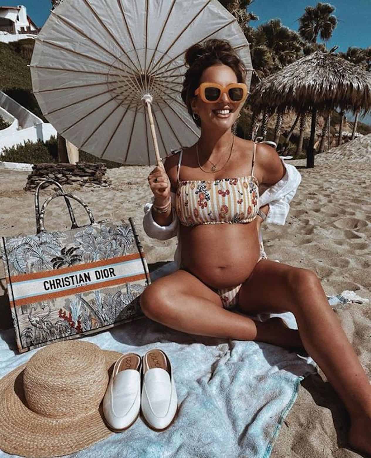 image of a pregnant woman sitting on a towel on the beach with an umbrella in her hand and a designer Dior bag at her side.