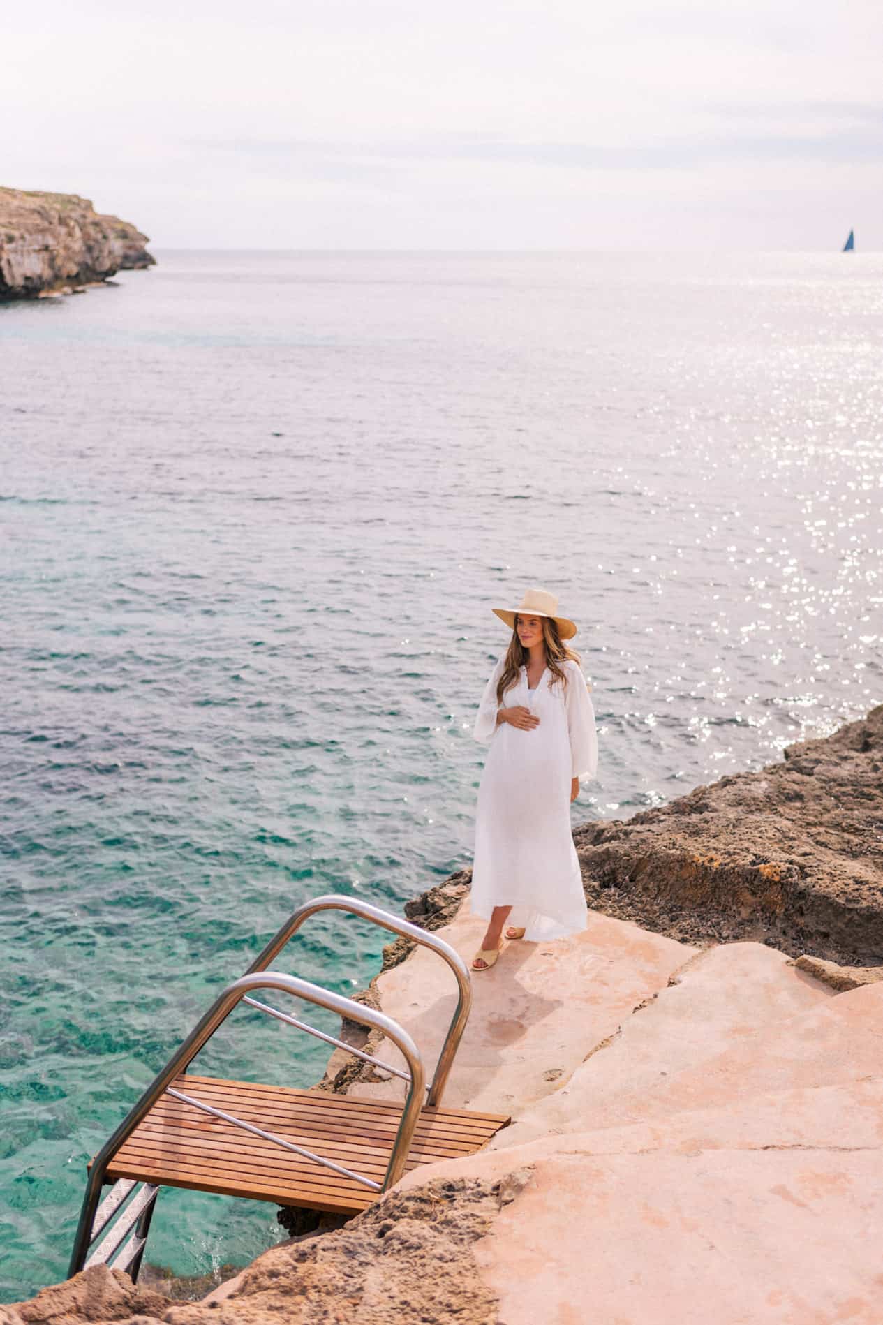 image of a woman from a distance standing in front of an ocean cliff wearing a white kaftan and a sun hat, she is pregnant