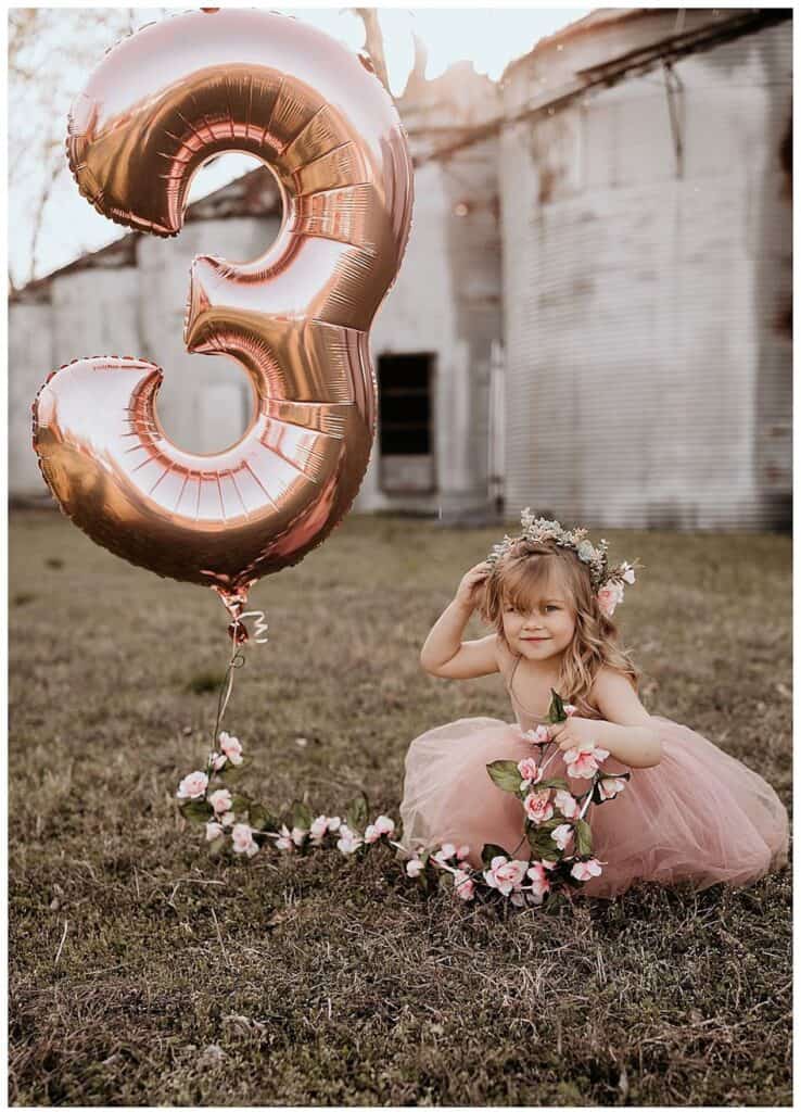 an image of a small girl sitting in a field in a pink dress holding a balloon that is the number 3