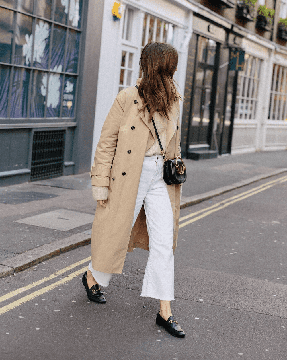 Woman wearing white jeans, black loafers, a cream sweater and a tan trench coat crosses the street.