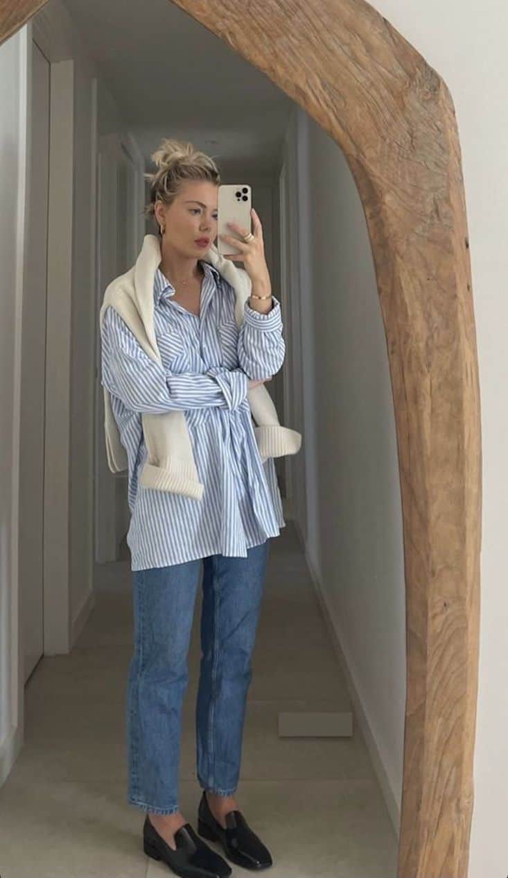 Woman looking in a mirror wearing jeans, a blue striped button down, black loafers, and a cream sweater over her shoulders.