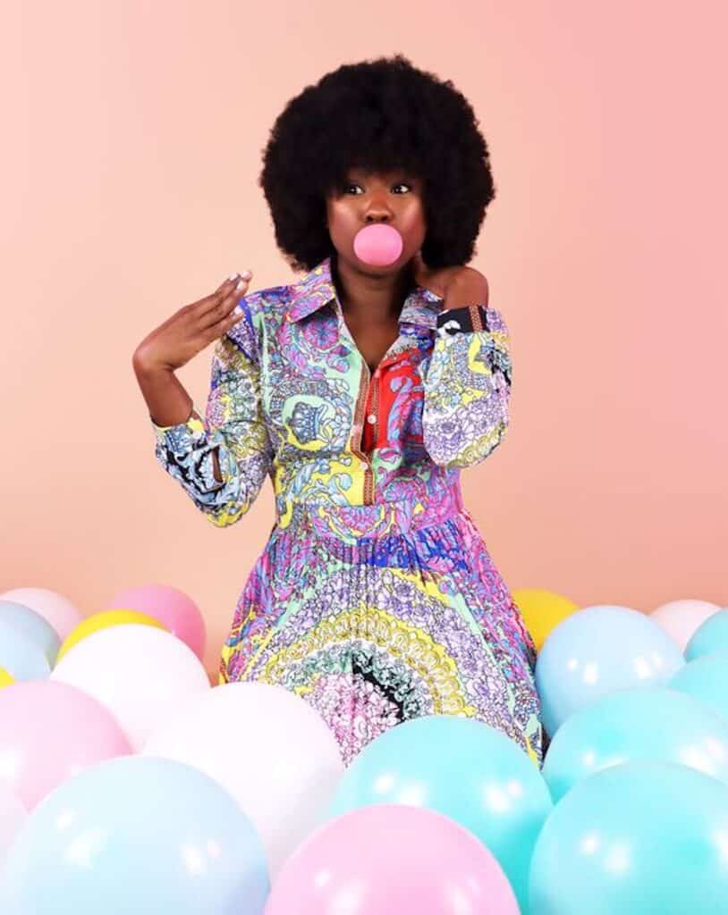 an image of a black woman sitting in a bed of balloons blowing a pink bubble gum bubble