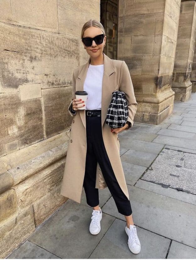 Woman wearing a tan trench coat, black jeans, white sneakers and a white t-shirt sips coffee in the city.