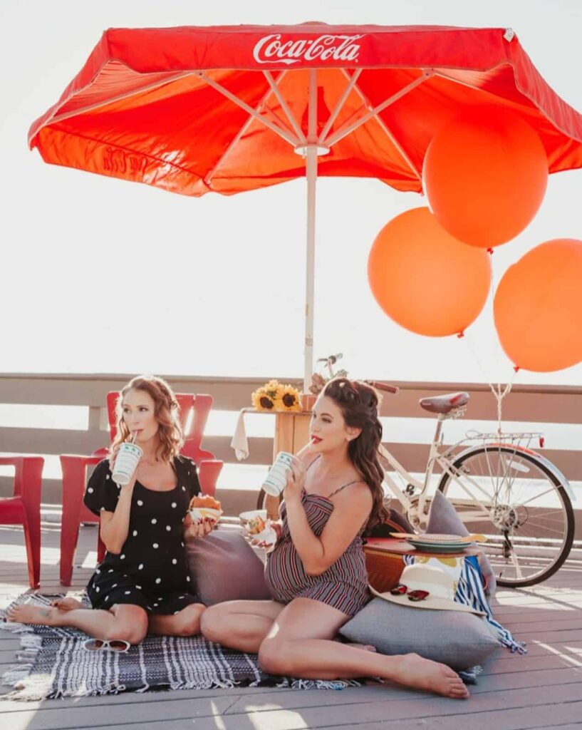 image of two women sitting on a pier, both pregnant, dressed in retro clothing and an orange umbrella and balloons behind them