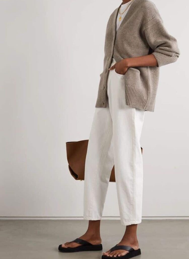 Woman wearing white jeans, black sandals, and grey sweater poses in front of a white wall.