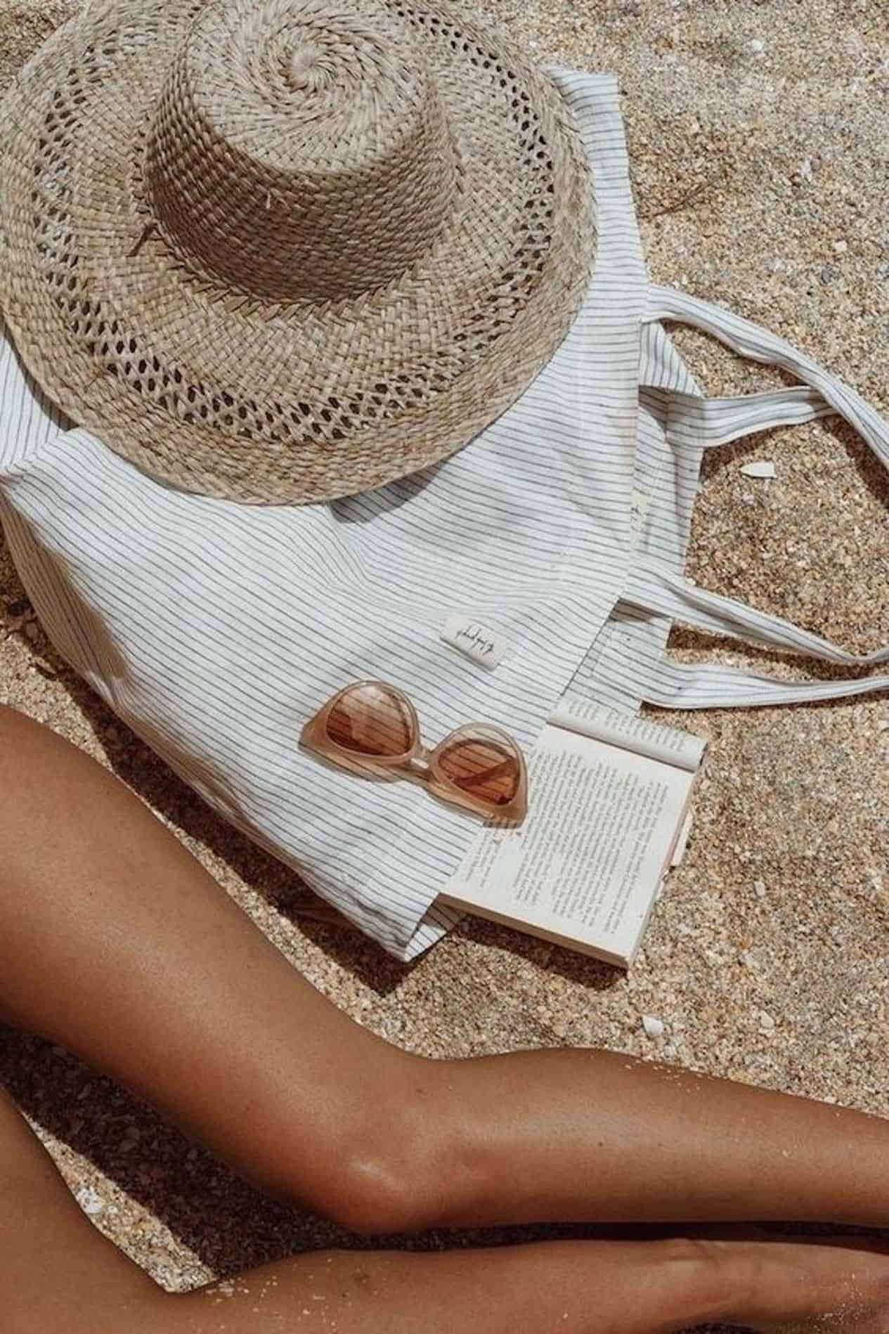 A women's legs, beach bag, sunglasses and sunhat laying on the sand.