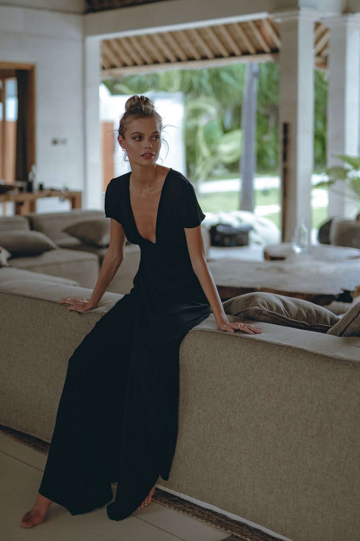 A woman in a black maxi dress leaning against a couch in a tropical destination.