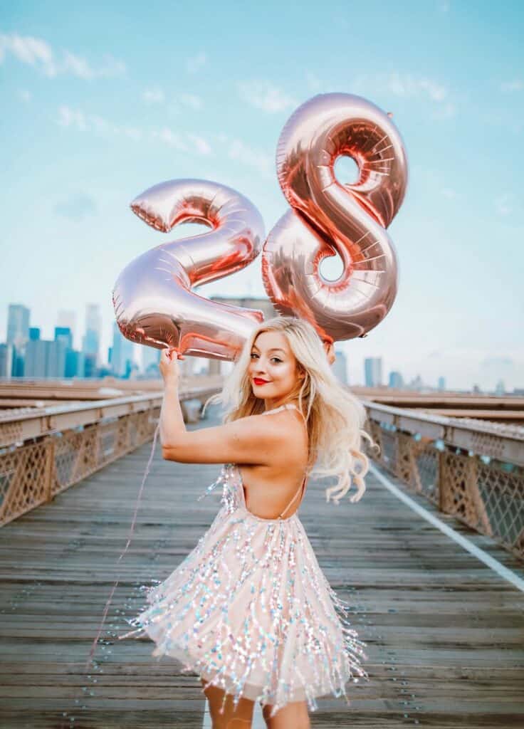 an image of a young blonde woman walking along a bridge holding two balloons that say "28"